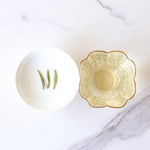 A white bowl with three green tea leaves sits next to a decorative glass cup filled with My Store's Zhu Ye Qing Green Tea, displayed on a white marble surface.