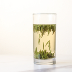 A glass of light-colored liquid with Zhu Ye Qing Green Tea leaves from My Store floating and settled at the bottom, displayed on a white surface, highlights this premium Chinese tea against a plain background.
